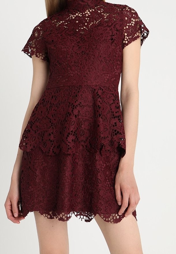 Femme portant une robe en dentelle bordeaux à manches courtes avec des motifs floraux et une jupe à volants sur un fond uni.