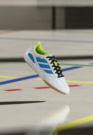 White indoor sports shoe with blue stripes, black laces, and neon green inner lining, hovering above a gym floor with colorful court lines.