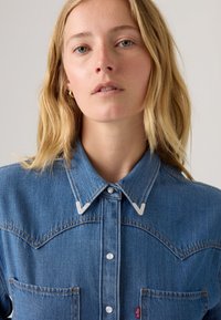 Jeanshemd mit klassischem Western-Design, spitzem Kragen, silbernen Nähakzenten und zwei Brusttaschen. In blauer Farbe.