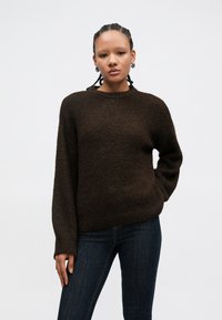 Bruine gebreide trui met een ronde hals, lange mouwen en een losse pasvorm. De textuur is zacht en licht pluizig. Draagt bij een donkere spijkerbroek.