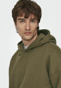 Felpa verde realizzata in tessuto morbido, con cappuccio regolabile e vestibilità comoda. La texture è liscia con dettagli di cucitura visibili.