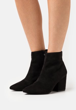 Bottines en daim noir avec un bout pointu, une fermeture éclair et un talon carré. Texture lisse et design ajusté couvrant la cheville.