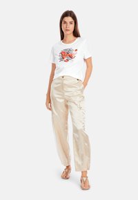 Weißes T-Shirt mit einem bunten Grafikdesign, kombiniert mit glänzenden beigen Cargo-Hosen. Das Outfit wird durch Riemchensandalen ergänzt.