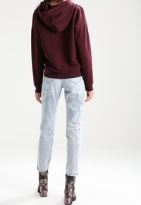 Sudadera burdeos con un bolsillo en la parte posterior, pantalones vaqueros desgastados de un azul claro y botas de tobillo con patrón texturizado en colores oscuros.