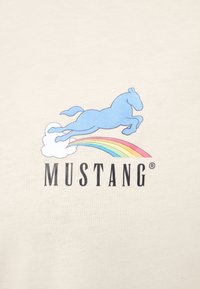 Cremfarbener T-Shirt mit einem blauen Pferd, das über eine Wolke mit einem Regenbogen springt, begleitet von dem Text "MUSTANG" in fetter schwarzer Schrift.