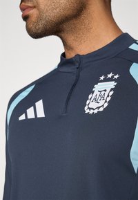 Ναυτικό αθλητικό μπουφάν με ανοιχτό μπλε λεπτομέρειες, γιακαδάκι quarter-zip και λογότυπο της Ομοσπονδίας Ποδοσφαίρου Αργεντινής. Η υφή φαίνεται λεία.