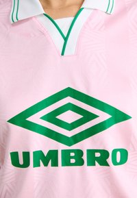 Camisola desportiva rosa claro com colarinho branco, detalhes verdes e um grande logótipo de diamante verde. O tecido apresenta uma suave textura geométrica.