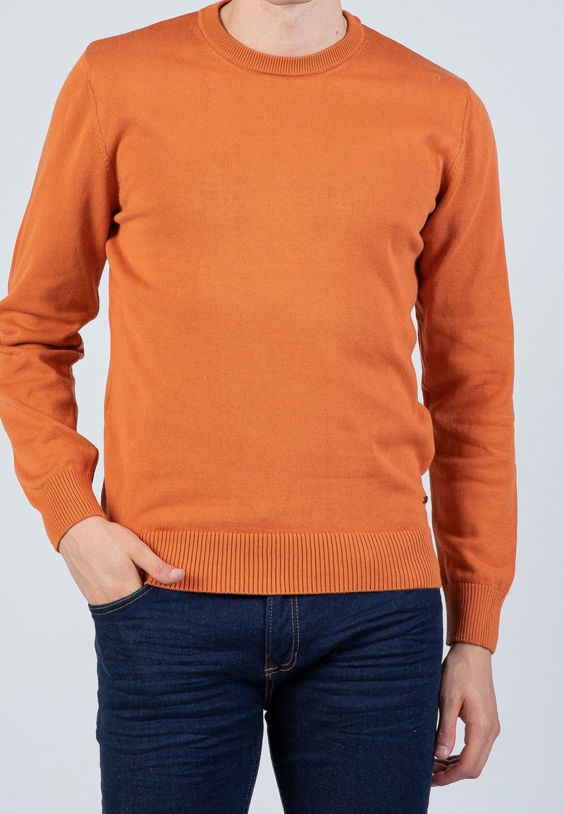 Homme portant un pull orange à manches longues et col rond avec un jean bleu foncé, une main dans la poche, sur un fond clair uni.