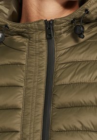 Veste matelassée verte avec une texture matelassée horizontale, une fermeture éclair noire et des cordons ajustables sur la capuche pour un ajustement parfait.