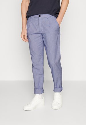 Homme portant un pantalon bleu clair avec revers, des baskets blanches et un t-shirt noir, debout avec une main dans la poche devant un fond uni.