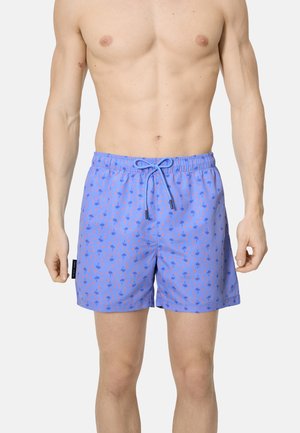 Hombre con pantalones cortos de baño azules con patrones de palmeras y estrellas en naranja y azul marino, de pie contra un fondo liso.