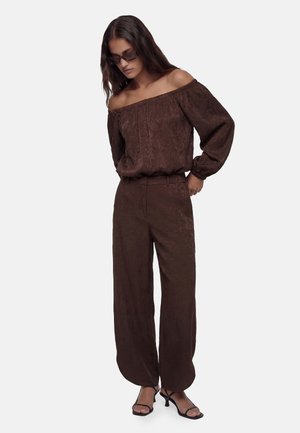 Kobieta w brązowej bluzce odsłaniającej ramiona, dopasowanych szerokich spodniach, czarnych sandałkach na obcasie i okularach przeciwsłonecznych, stojąca z rękami w kieszeniach.