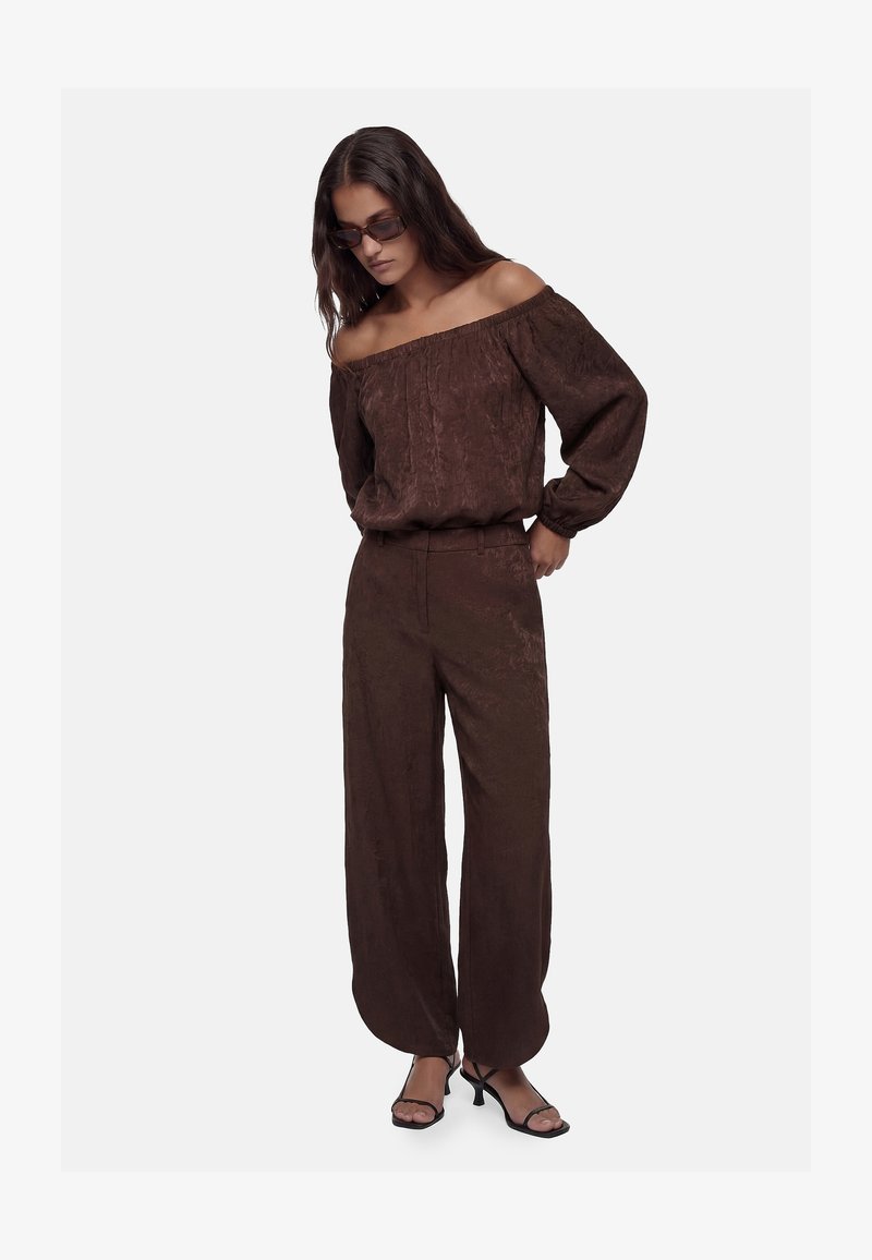 Mujer con blusa marrón sin hombros, pantalones anchos a juego, sandalias negras de tacón y gafas de sol, de pie con las manos en los bolsillos.