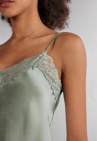 Satin camisole in zachte groene kleur met delicate kantafwerking langs de halslijn en verstelbare spaghettibandjes. Glad textuur, aansluitend ontwerp.