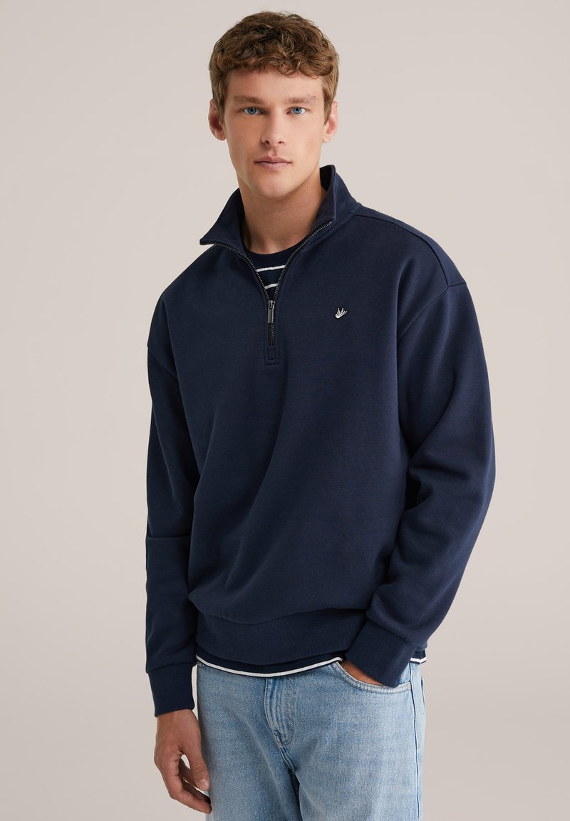 Tengerészkék pulóver fél cipzáras gallérral, puha tapintású és bordázott mandzsettákkal. Kis logó található a mellrészen, csíkos inggel rétegezve.