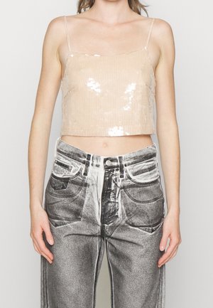 Woman wearing a beige sequin crop top with thin straps and high-waisted, black-and-white acid-wash jeans, standing against a plain background.