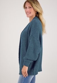 Overmaatse teal cardigan van gestructureerde brei-stof, met een diepe V-hals en geribbelde manchetten, gedragen over een blauwe jeans.
