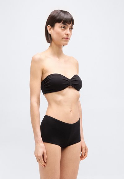 Γυναίκα με κοντά σκούρα μαλλιά που φοράει μαύρο μπικίνι χωρίς τιράντες και μαγιό ψηλής μέσης, στέκεται μπροστά σε ένα απλό ανοιχτό φόντο.