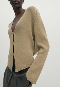 Frau in beiger Strickjacke mit tiefem V-Ausschnitt und schwarzen Lederhosen, die mit einer Hand in der Tasche vor einem schlichten Hintergrund posiert.