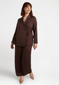 Brązowy, lekki zestaw odzieży domowej z topem w stylu kopertowym z wiązaniem w talii i spodniami o szerokich nogawkach. Całość dopełniają błyszczące obcasy.