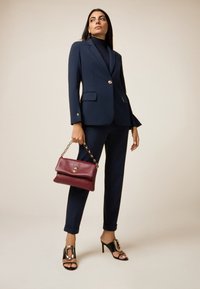 Completo giacca e pantaloni blu navy con un solo bottone, abbinato a una borsa borgogna e sandali neri open-toe con dettagli dorati.