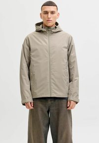 Veste beige clair imperméable avec capuche, fermeture éclair devant et cordons de serrage réglables. Texture lisse, coupe décontractée et manches longues.