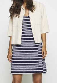 Chemise beige à manches courtes boutonnée par-dessus une robe rayée bleu marine et blanc. La robe présente une fente latérale et une coupe décontractée.