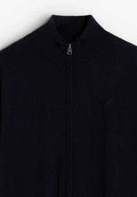 Suéter de cremallera azul marino oscuro con cuello alto, hecho de tejido de punto suave. Cuenta con una cremallera metálica y un sutil logo de calavera en el pecho izquierdo.