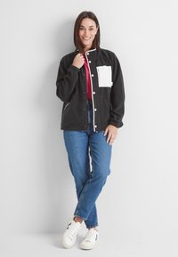 Black fleece jacket with white chest pocket, snap buttons, and elastic cuffs. Paired with blue jeans and white sneakers. Smooth texture.