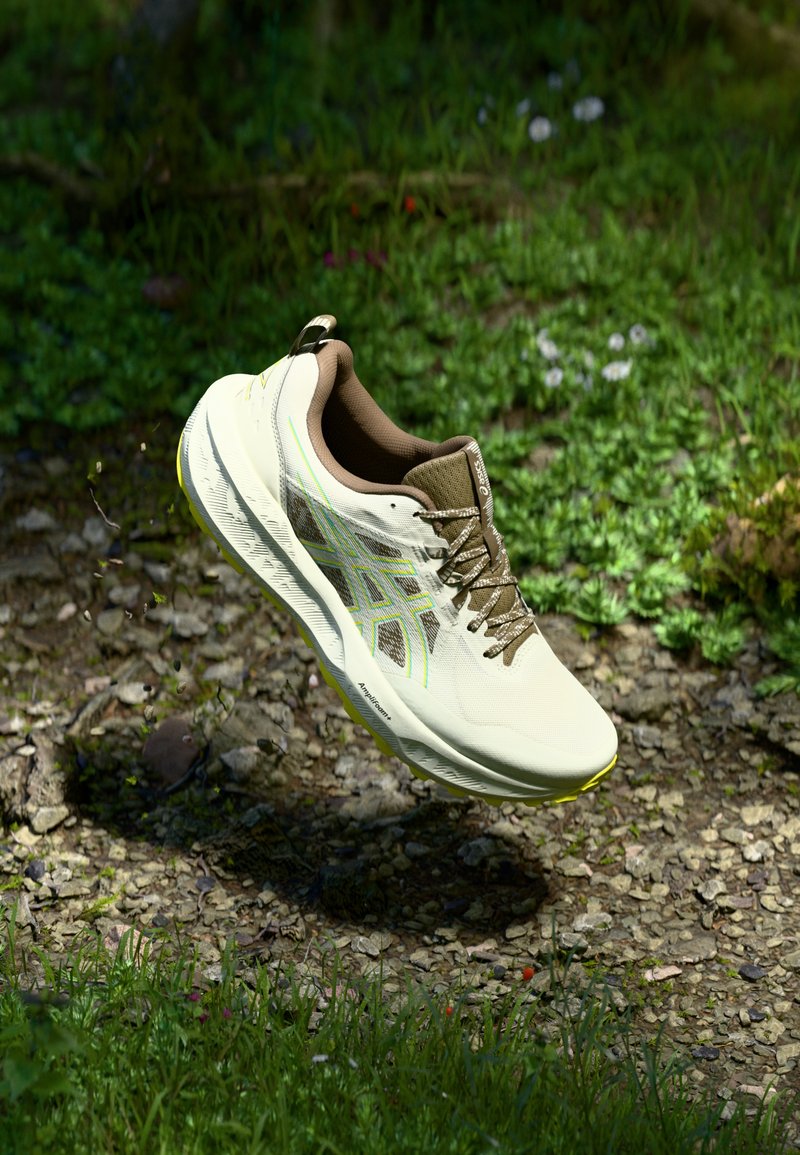 Helle beigen Sportschuh mit strukturiertem Obermaterial, braunen Akzenten und einem reflektierenden Design. Verfügt über eine gepolsterte Sohle und Schnürsenkel.