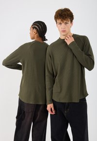 Olīvzaļās garās piedurknes krekli ar ribotu tekstūru, apaļiem kakla izgriezumiem un raglana piedurknēm; apvienoti ar tumšiem vaļīgiem biksēm ar kabatām.