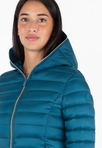 Giacca puffer turchese con chiusura a zip, cappuccio aderente e cuciture orizzontali. Include un'etichetta con logo sulla spalla sinistra. Tessuto morbido e liscio.