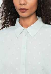 Light blue button-up shirt with a point collar, featuring raised polka dot patterns and a textured fabric. Close-up view of the neckline.