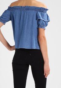 Top in denim a spalla scoperta con maniche corte a sbuffo, scollatura arricciata e design corto. Tessuto azzurro chiaro con una texture liscia.