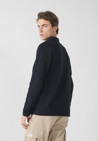 Polo shirt nero a maniche lunghe con colletto, dotato di fessure laterali e una texture liscia. Indossato con pantaloni cargo beige.