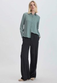Camicia verde chiaro abbottonata abbinata a pantaloni neri larghi. Il tessuto sembra morbido, con una superficie liscia e una silhouette rilassata.