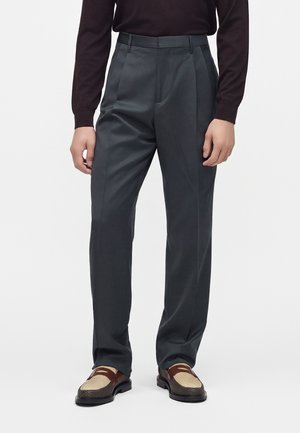 Dunkelgraue, plissierte Herrenhose kombiniert mit einem schwarzen Langarmtop und braunen sowie beigen Lederloafers vor weißem Hintergrund.