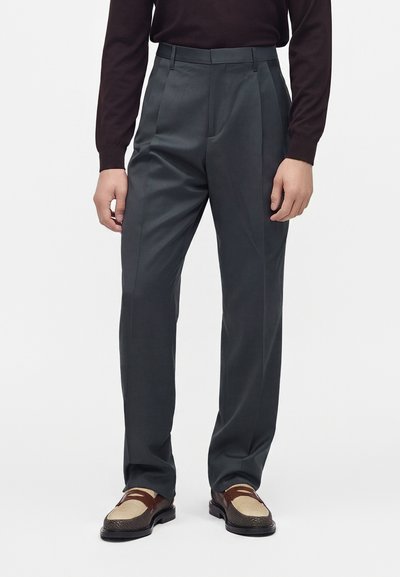 Pantalon plissé gris foncé pour homme, associé à un haut noir à manches longues et des mocassins en cuir marron et beige sur fond blanc.