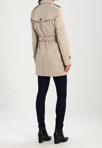 Henkilö, jolla on beige vyöllinen trenssi, tummat kapeat farkut ja mustat korolliset nilkkabootsit, seisoo valkoista taustaa vasten.