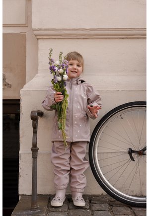 Világos rózsaszín vízálló kabát kapucnival és virágos díszítéssel, hozzáillő nadrág, virágokat tartva, egy kerékpár mellett, egy texturált fal előtt.