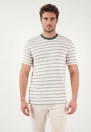 Homme portant un t-shirt blanc à manches courtes avec de fines rayures horizontales grises et un pantalon de couleur claire, debout devant un fond uni.