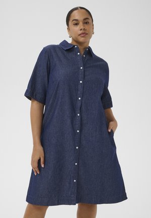 Nő térd alá érő, rövid ujjú, sötétkék farmer, galléros, gombos ruhában, oldalzsebekkel, egyszínű háttér előtt.