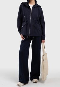 Navy windbreaker jacket with a hood, drawstring waist, and two front pockets, paired with dark wide-leg pants and a light beige tote bag.