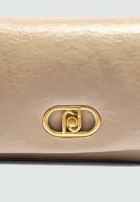 Zmačkaná béžová kožená kabelka s výrazným zlatým logem. Hladký povrch a minimalistický design, prezentující měkký obdélníkový tvar.