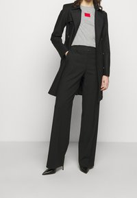 Abrigo negro sobre una camiseta gris con un logo rojo, combinado con pantalones negros de pierna ancha y tacones negros puntiagudos. Texturas de tela suaves.