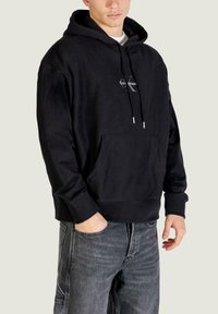 Sudadera negra con capucha hecha de tela suave, con un bolsillo frontal, capucha con cordón y un pequeño logo en el pecho. Combinada con jeans grises.