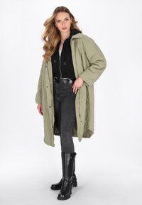 Olivengrøn quiltet frakke over en sort hættetrøje og grå skinny jeans. Sorte knæhøje støvler med klodsede hæle og sølv hardware detaljer.