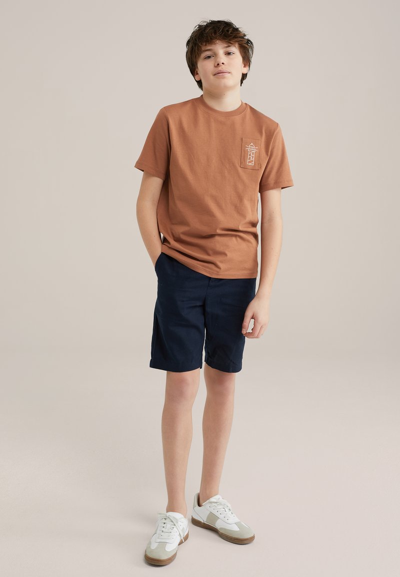 Brun bomull T-shirt med fickgrafik, matchad med marinblå shorts och vita sneakers med gråa accenter. Enkel design, avslappnad passform.