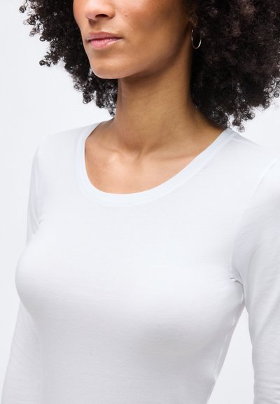 Γυναίκα με σγουρά μαλλιά που φοράει λευκό πουκάμισο με μακριά μανίκια και ασημένιο κρίκο στο αυτί, εμφανιζόμενη από τους ώμους έως το πηγούνι σε λευκό φόντο.