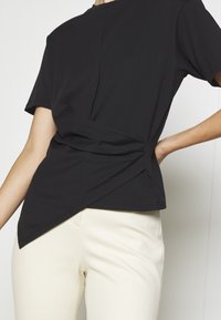 T-shirt en coton noir avec un design à l'avant torsadé, manches courtes et coupe décontractée, associé à un pantalon crème texturé.
